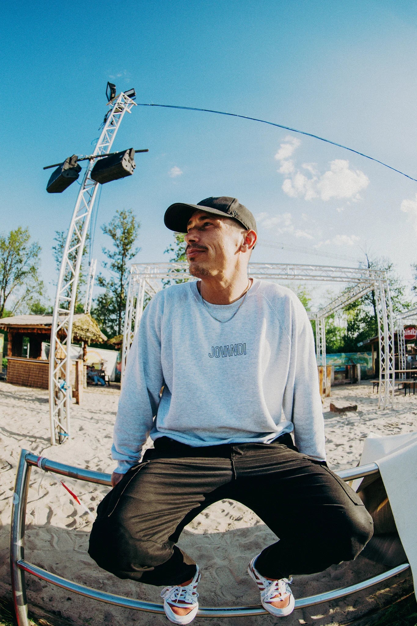 Man with a heather grey Lines Sweater is sitting on a fence in the skatepark with beach in the background on a sunny day