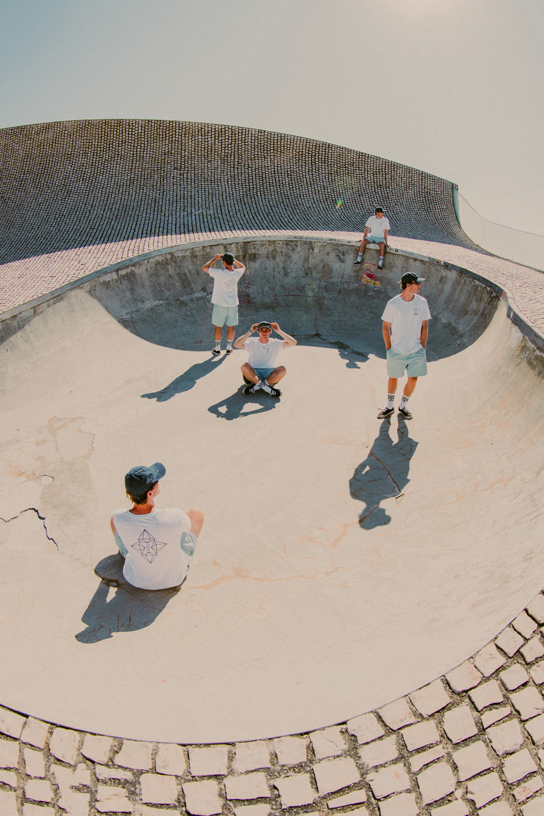 Model wearing Jovandi Diamonds T-Shirt, light blue shorts, blue cap and sunglasses, cloned multiple times in a rooftop bowl in Biarritz, France under a bright blue summer sky.